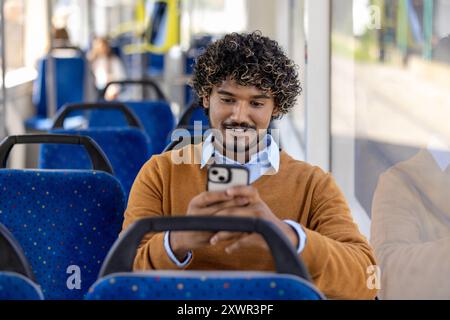 Mann, der im Bus sitzt und ein Smartphone benutzt, mit einem Lächeln. Zwangloses Ambiente in öffentlichen Verkehrsmitteln mit blauen Sitzen und Fensterbeleuchtung. Motivierte und entspannte Momente festgehalten. Stockfoto