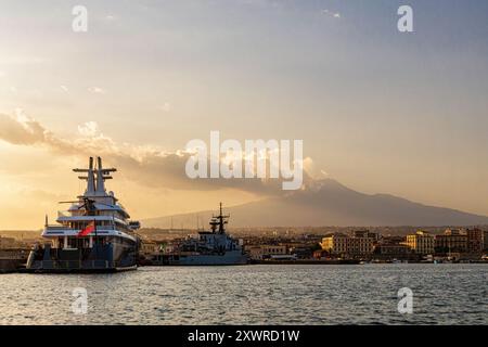Malerischer Blick auf den Hafen von Catania bei Sonnenuntergang mit Luxusyacht, Militärschiff und dem Ätna im Hintergrund. Goldene Stunde Licht über der sizilianischen Küste, Stockfoto