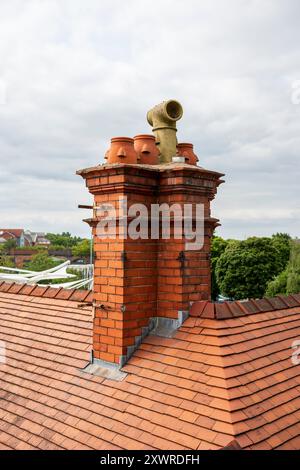 Alter roter Backsteinkamin auf einem gekachelten Dach, umgeben von üppigen grünen Bäumen und bewölktem Himmel im Hintergrund. UK Stockfoto