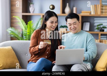 Glückliche asiatische Familie reifes Paar, das auf der Couch sitzt und mit Laptop und Kreditkarte online einkaufen kann. Sowohl lächelnd als auch aktiv, zeigt Lo Stockfoto