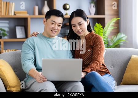 Asiatisches reifes Ehepaar genießt eine gute Zeit zu Hause, sitzt auf dem Sofa und benutzt zusammen einen Laptop. Lächelnd und entspannt, durchstöbern und verbinden sie sich und zeigen M Stockfoto