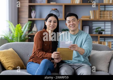 Asiatisches Paar sitzt bequem auf dem Sofa und lächelt zusammen, während er den Umschlag hält. Sie strahlen Wärme und Glück in gemütlicher Wohnzimmeratmosphäre aus und zeigen starke Beziehungen und Kommunikation. Stockfoto