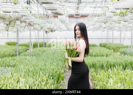 Eine junge Frau, die in einem großen Gewächshaus steht und eine blühende Tulpe in den Händen hält. Stockfoto