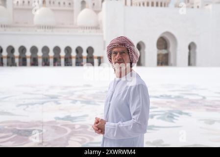 Ein Mann in Kanroa-traditioneller arabischer Kleidung steht vor der Scheich-Zayed-Moschee in Abu Dhabi mit majestätischen Kuppeln und Bögen im Hintergrund, cr Stockfoto