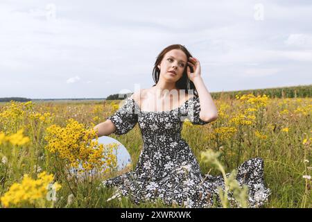 Eine junge Frau in schwarz-weißem Kleid, die zwischen gelben Wildblumen auf einem Feld unter bewölktem Himmel sitzt und einen Spiegel und einen Blumenstrauß hält Stockfoto