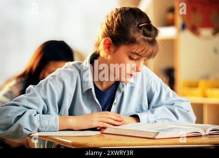 Eine junge Studentin konzentriert sich auf ihr Lehrbuch, die in das Studium eingetaucht ist, während sie an einem Schreibtisch in einem hellen Klassenzimmer sitzt. Stockfoto