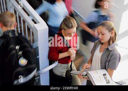 Zwei Mittelschüler diskutieren mit einem Lehrbuch über die Schularbeit, während andere Schüler in einer Schule durch den belebten Flur ziehen. Stockfoto