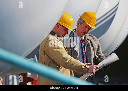 Ingenieure analysieren Blaupausen, während sie in der Nähe großer Industrieanlagen stehen, und konzentrieren sich dabei auf Projektdetails und Sicherheitsmaßnahmen. Stockfoto