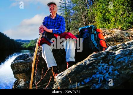 Mit seinem Rucksack an seiner Seite sitzt Ein älterer Mann auf einem großen Felsen am Fluss, hält einen Wanderstock und genießt die friedliche Waldlandschaft Stockfoto