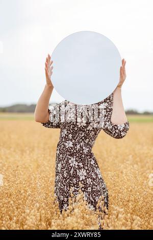 Naturbegriff, geisteszustände und geheimnisvolles Konzept. Eine Frau, die einen Spiegel vor dem Kopf hält, der den Himmel reflektiert, in einem goldenen Weizen gefallen Stockfoto