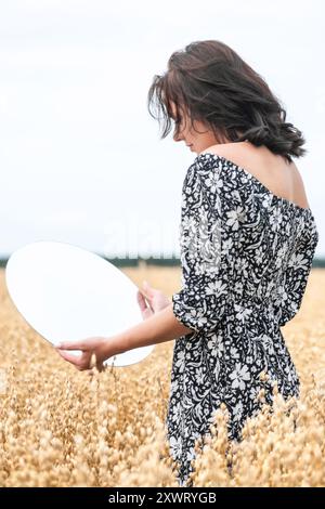Eine junge Frau in einem Blumenkleid hält einen runden Spiegel in einem goldenen Weizenfeld. Das Bild enthält Rauschen und Bewegungsunschärfe. Selektiver Fokus Stockfoto