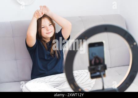 Kleines Mädchen, das vor dem Smartphone auf einem grauen Sofa in einem hellen Raum einen Videoblog aufnimmt. Ein Mädchen in einem blauen T-Shirt zeigt vorne einen Häschen Stockfoto