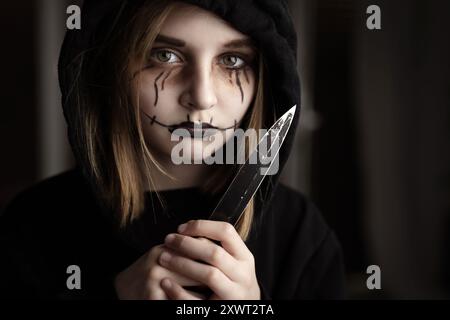 Ein gruseliges kleines Mädchen, das ein Messer hält und die Klinge ansieht. Gruseliges Kind in schwarz und Halloween dunkel Make-up Stockfoto