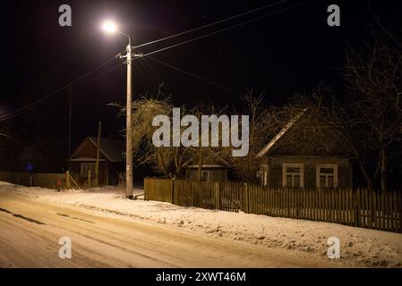 Eine friedliche Dorfstraße, bedeckt mit Schnee, in der Nacht in das warme Licht einer Straßenlaterne getaucht. Die Szene fängt die Ruhe und Stille einer Winternacht in der Nähe des Nationalparks Białowieża ein. Stockfoto