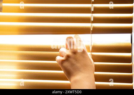 Die Hand eines kleinen Jungen öffnet gelbe Jalousien an einem Fenster in einer Wohnung Stockfoto