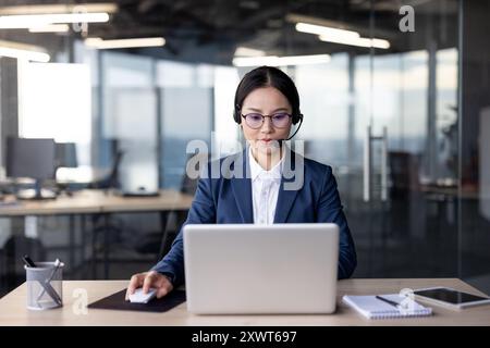 Asiatische Geschäftsfrau mit Brille und Headset, die im Büro an einem Laptop arbeitet, um Produktivität zu erzielen Stockfoto