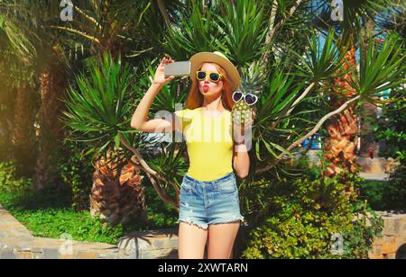 Porträt der schönen jungen Frau, die Selfie mit Smartphone und Ananas in der Sonnenbrille im Sommerpark auf Palmenhintergrund macht Stockfoto