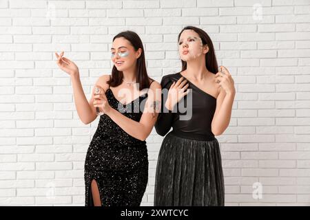 Schöne junge Frauen mit Gesichtsmaske und Flecken unter den Augen, die Flaschen mit Parfüm in der Nähe der weißen Ziegelwand auftragen Stockfoto