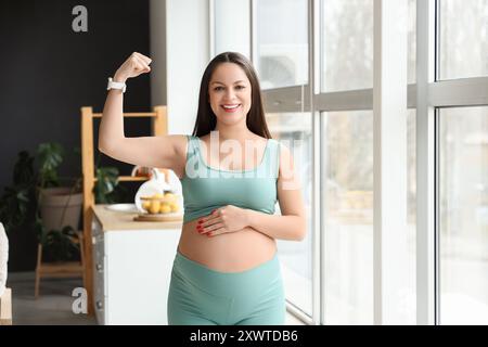 Sportliche schwangere Frau, die zu Hause Muskeln in der Nähe des Fensters zeigt Stockfoto