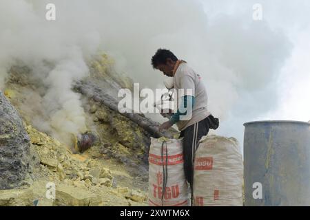 Ein Mann verpackt abgebauten Schwefel am Kratersee des Iljen für die Träger. Eingekreist von einer zerfurchten Felswand liegt im Krater des Gunung Iljen der Kratersee. Seine grünlich-blaue Farbe ist auf hohe Konzentrationen von Alaun, Schwefel und Gips zurückzuführen. Mit einem pH-Wert um 0,3 und seiner wird er als größtes Säurefass der Erde bezeichnet. Kratersee - Breite: 600 m - Länge: 960 m - Tiefe: bis zu 200 m *** Ein Mann packt am Kratersee Iljen für die Träger eingekreist von einer gefurchten Felswand liegt der Kratersee im Krater von Gunung Iljen in seiner grünlich-blauen Farbe Stockfoto