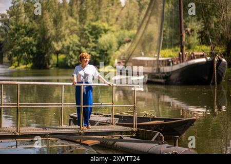 Eine Frau genießt die ruhige Atmosphäre am Fluss, die auf einem Dock mit einem rustikalen Boot im Hintergrund unter klarem Himmel steht. Stockfoto