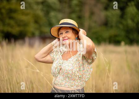 Eine fröhliche Frau steht auf einem Feld, justiert ihren Hut und lächelt, umgeben von hohem Gras und sanftem Sonnenlicht. Stockfoto