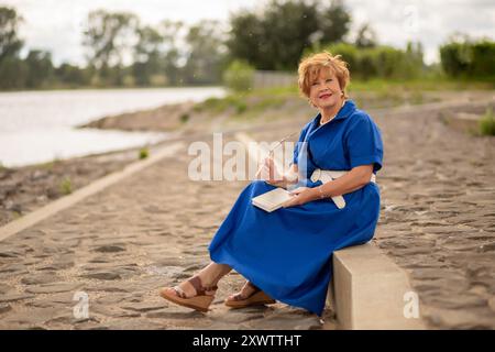 Eine Frau genießt einen friedlichen Moment am Wasser und schreibt Gedanken in ihr Notizbuch, während sie auf einem Steinweg sitzt. Stockfoto