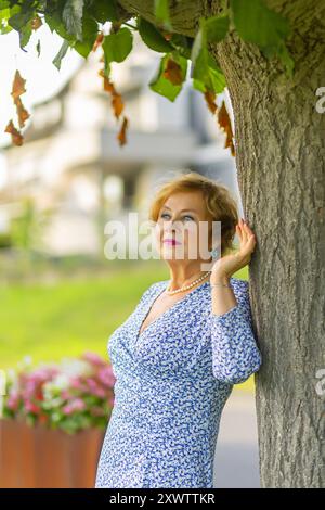 Eine Frau steht an einem Baum und genießt den Sonnenschein, trägt ein blau gemustertes Kleid und trägt elegante Ohrringe. Stockfoto