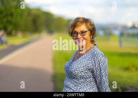 Eine Frau mit Brille steht souverän in lässiger Kleidung und genießt einen sonnigen Tag in einem lebhaften Park. Stockfoto
