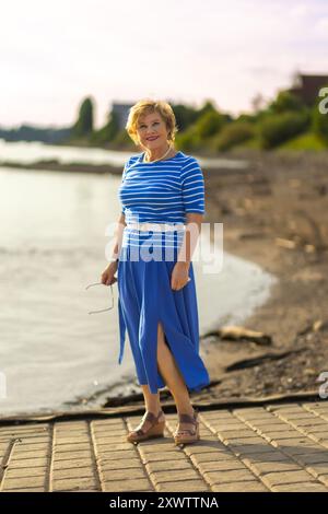 Eine Frau posiert anmutig am Wasserrand, trägt ein blau-weiß gestreiftes Kleid und genießt den warmen Tag. Stockfoto