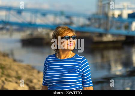 Eine Frau in einem blau-weiß gestreiften Hemd genießt das sonnige Wetter, während sie am Wasser steht und selbstbewusst mit einer Sonnenbrille lächelt. Stockfoto