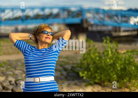 Eine Frau, die sich draußen am Wasser entspannt und sich im Sonnenlicht sonnt, während sie eine Sonnenbrille und ein stilvolles gestreiftes Hemd trägt. Stockfoto