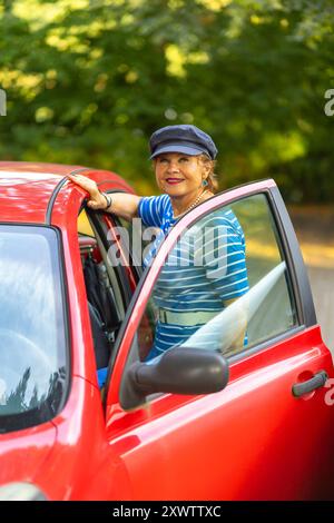 Eine fröhliche Frau lehnt sich mit einem gestreiften Hemd und einem blauen Hut an ein rotes Auto mit landschaftlich schöner grüner Kulisse. Stockfoto