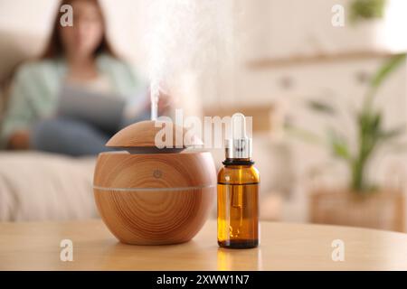 Aromadiffusor und Flasche ätherisches Öl auf dem Tisch, Platz für Text. Frau liest Buch im Zimmer, selektiver Fokus Stockfoto