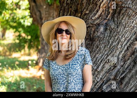 Porträt einer älteren weißen Frau, die Kleid und Hut neben dem Stamm eines riesigen Baumes auf dem Feld trägt. Stockfoto