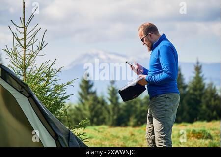 Der Tourist steht neben dem Zelt, benutzt ein Smartphone und hält ein tragbares Solarmodul-Ladegerät. Schneebedeckte Berge und üppig grüne Bäume, Harmonie von Technologie und Natur in malerischer Umgebung im Freien. Stockfoto