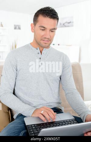 Der Mann saß mit überkreuzten Beinen auf dem Sofa und benutzte einen Laptop Stockfoto