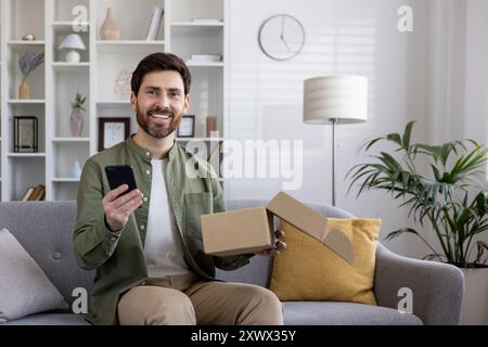 Lächelnder Mann, der auf der Couch sitzt, Smartphone hält und Pappkarton öffnet und Glück und Zufriedenheit zeigt. Die Szene veranschaulicht Online-Shopping, Unboxing und Technologiekonzepte. Stockfoto