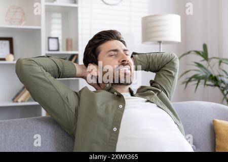 Mann in lässiger Kleidung, der Entspannung und Komfort auf dem Sofa erlebt. Hände hinter dem Kopf, die Ruhe und Glück zeigen. Helle und luftige Wohnzimmerumgebung, die ein Gefühl der Zufriedenheit fördert. Stockfoto