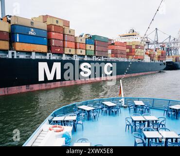 Blick auf ein großes Containerschiff mit bunten Frachtcontainern, das am belebten Containerhafen in Hamburg angedockt ist. Im Vordergrund stehen leere Liegestühle auf einem Schiff, die einen Kontrast zwischen Handel und Freizeit darstellen. Stockfoto