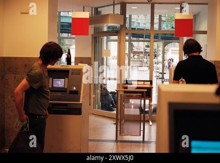 Eine Alltagsszene in einer Berliner Bankfiliale mit Leuten, die Geldautomaten benutzen. Das Bild fängt die tägliche Routine ein und deutet auf persönliche Momente und finanzielle Transaktionen hin, mit einem Kontrast zu den Straßen draußen. Stockfoto