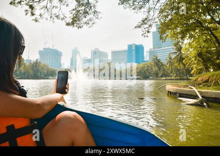 Junge kaukasische Frau genießt sonnigen Tag im Park auf Paddelbootfilm mit Smartphone wilde Riesenechse. Besuchen Sie den öffentlichen Lumpini Park, den beliebtesten Park in Stockfoto