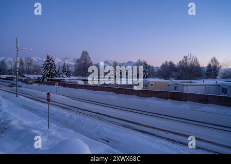 Eine ruhige Winterszene bei Sonnenaufgang mit einem schneebedeckten Wohnwagenpark in Anchorage, Alaska. Die ruhige Wohngegend wird von hohen Bergen und schneebedeckten Bäumen eingerahmt und verkörpert die Ruhe des Winters. Stockfoto