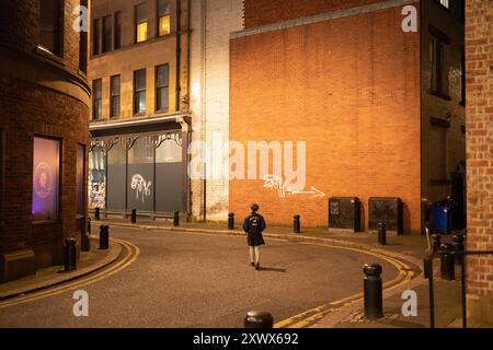 Eine Einzelfigur läuft nachts in einer Gasse in Newcastle upon Tyne. Die Stadtlampen strahlen ein warmes Leuchten auf die urbane Umgebung aus. Die Szene weckt Gefühle der Einsamkeit und Erkundung inmitten der mit Graffiti bedeckten Wände. Stockfoto