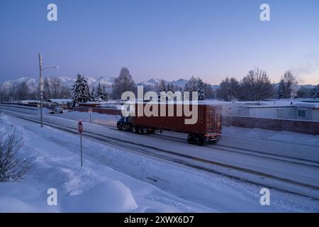 Landschaftlich reizvoller Wintermorgen in einem Wohnwagenpark in Anchorage, Alaska, mit einem Sattelzug, der auf einer verschneiten Straße vorbeifährt. Schneebedeckte Bäume und ferne Berge schaffen eine ruhige, kalte Atmosphäre. Stockfoto