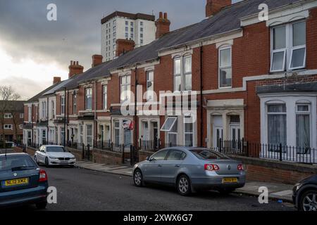 Eine typische Wohnstraße in Fenham, Newcastle upon Tyne, gesäumt von Reihenhäusern und geparkten Autos unter bedecktem Himmel, die das städtische Leben einfangen. Die Szene symbolisiert Gemütlichkeit trotz des trostlosen Wetters. Stockfoto