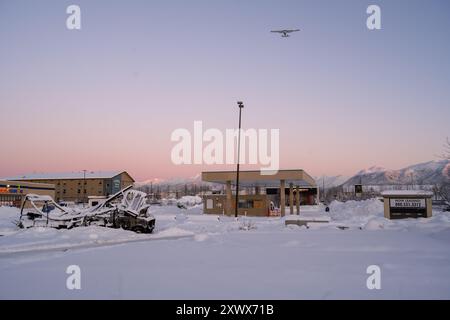 Eine verschneite Winterlandschaft in der Nähe des Merrill Field Airport in Anchorage, Alaska. Das Bild zeigt ein kleines Flugzeug, das über dem Himmel fliegt, einen schneebedeckten Parkplatz und verschiedene Gebäude unter einem pastellfarbenen Himmel. Stockfoto