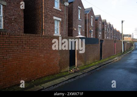 Eine enge Gasse verläuft an einem klaren Tag in Fenham, Newcastle upon Tyne, entlang einer Reihe von Ziegelterrassenhäusern mit einer hohen Ziegelmauer. Das Bild kann Uniformität und Grenzen symbolisieren. Stockfoto