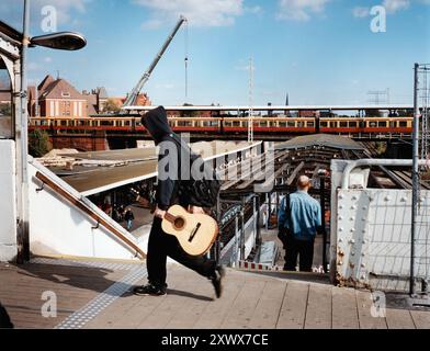 Ein Mann läuft mit einer Gitarre im Bahnhof Ostkreuz in Berlin im Jahr 2005. Das Bild erfasst eine sich wandelnde Stadtlandschaft mit Zug und Kran und symbolisiert den Schnittpunkt von Fahrten und Umwandlungen. Stockfoto