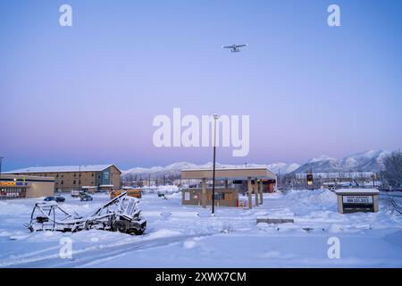 Ein malerischer Winterblick mit schneebedeckten Gebäuden und einem Flugzeug, das in der Nähe des Merrill Field Airport mit majestätischen Bergen im Hintergrund fliegt, Anchorage, Alaska. Dieses Bild erinnert an Themen der Ruhe und Widerstandsfähigkeit. Stockfoto